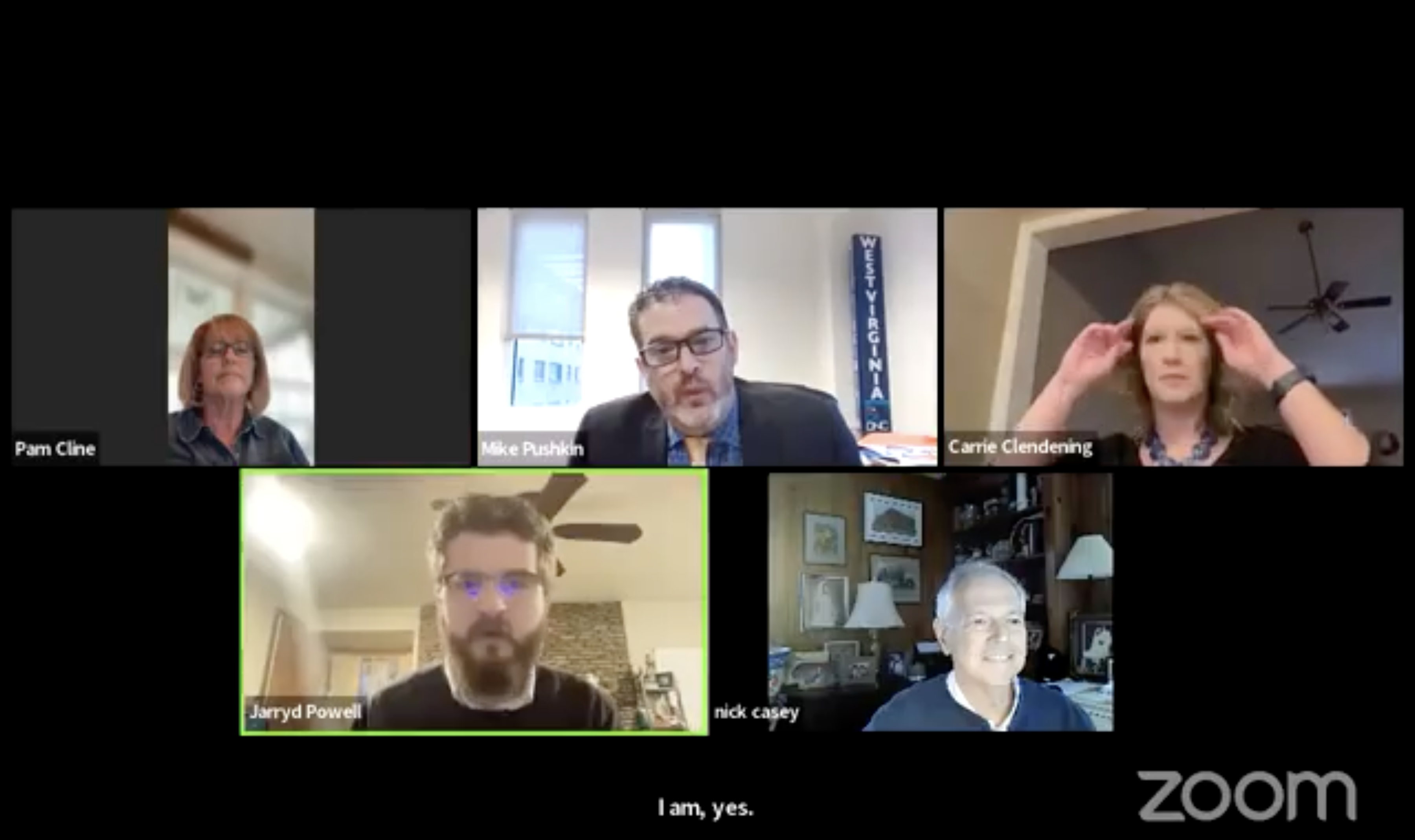 A screengrab from a Zoom video conference meeting. The black screen is divided into five video windows arranged in a 3-2 grid.  The top middle window, which is slightly prominent, features Delegate Mike Pushkin, who is actively speaking. He is a Caucasian man with glasses and dark hair, wearing a dark blazer over a multi-colored shirt and tie. In his background, a tall banner reads "WEST VIRGINIA" vertically with a "DNC" (Democratic National Committee) logo below it.  The top left window features a woman labeled "Pam Cline." She is Caucasian, wearing glasses, a denim jacket, and a necklace. The top right window features a Caucasian woman named "Carrie Clendening," who is adjusting her reddish-brown hair with both hands.  The bottom left window features a man named "Jarryd Powell," who has dark hair, glasses, and a full beard. His window is highlighted with a green border, indicating he may be speaking or was recently active. The bottom right window features an older Caucasian man with white hair named "nick casey" (all lowercase), who is smiling.  A large white "zoom" logo is visible in the bottom right corner of the entire screengrab. At the bottom, centered in the black background, are the captions: "I am, yes."